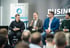 Vier Männer sitzen auf einer Bühne, einer spricht ins Mikrofon vor einem Banner mit "BUSINESS.DE" und "PROZESS AGENTUR".