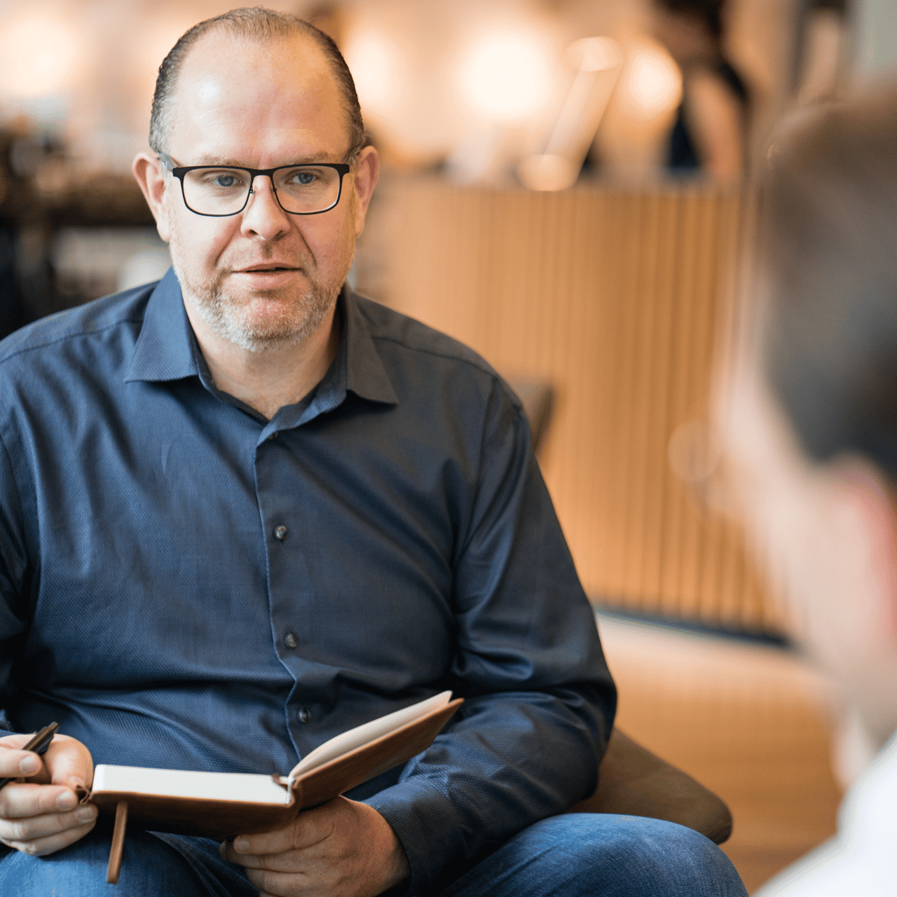 Ein Mann mit Brille und blauem Hemd sitzt in einem Gespräch und hält ein aufgeschlagenes Notizbuch.