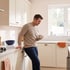 Man in a tan sweater leans against a white kitchen counter holding a mug with a pained facial expression.