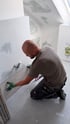 Handwerker kniet im Rohbau und bringt eine graue Fliese an einer weißen Wand mit Rohrleitungen an.