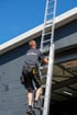 Norman Singer mit Werkzeuggürtel steigt an einer grauen Hauswand auf eine hohe Aluminiumleiter bei blauem Himmel.