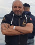 Ein kahler Mann mit Bart, schwarzem Kapuzenpullover und tätowiertem Arm verschränkt die Arme vor dem Wasser.