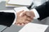 Zwei Personen in Anzügen schütteln sich die Hände über einem Schreibtisch mit Dokumenten.