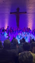 Chor in weißen Roben mit blauen Akzenten singt vor einem Kruzifix und Publikum in lila Licht.