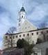 Weiße Kirche mit grünem Zwiebelturm und Uhr auf einem Hügel, umgeben von Gebäuden und kahlen Bäumen.