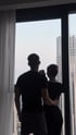 Silhouetted couple standing by a large window, looking out at a city skyline with tall buildings under a bright sky.