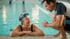 Ältere Frau mit blauer Schwimmbrille lächelt einen Trainer am Beckenrand eines Schwimmbads an.
