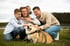 Eine lächelnde vierköpfige Familie hockt auf einer Wiese und streichelt ihren Corgi-Hund vor einer Waldkulisse.