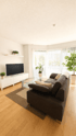 Modernes Wohnzimmer mit dunklem Sofa, Glastisch, Fernseher auf weißem Lowboard und Zimmerpflanzen vor hellem Fenster.