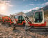 Eine Reihe von orange-weißen Takeuchi TB153R Baggern auf steinigem Gelände vor Bergen.