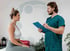 Female patient in sports bra talks to male chiropractor holding a clipboard and pen.