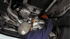 Mechaniker mit Baseballmütze, orangefarbenen Handschuhen und Lampe arbeitet unter einem Auto auf einer Hebebühne.