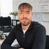 Lächelnder Mann mit Bart im Büro vor Plänen zu Leitungswasser, Feuchte und Wasserschäden mit Messtechnik im Hintergrund.