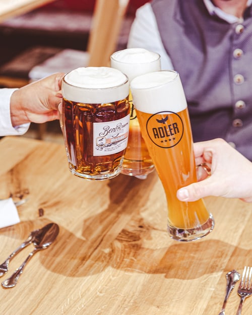 Drei Personen stoßen mit Biergläsern an, ein Glas mit Adler-Logo