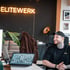 Mann mit Tattoos lacht am Schreibtisch; Leuchtreklame „ELITEWERK“ an schwarzer Wand.