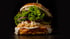 Ein großer Burger mit Salat, Zwiebeln, Gurken, Soße und zerbröseltem Feta auf dunklem Hintergrund.