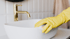 Hand mit gelbem Gummihandschuh reinigt Waschbecken mit Schwamm und Seifenblasen.