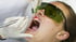 Eine Person mit Schutzbrille erhält eine zahnärztliche Laserbehandlung, wobei ein Instrument in den offenen Mund gehalten wird.