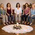 Fünf Frauen meditieren sitzend in einem Kreis um eine brennende Kerze und Blumenarrangement auf einem weißen Tuch.