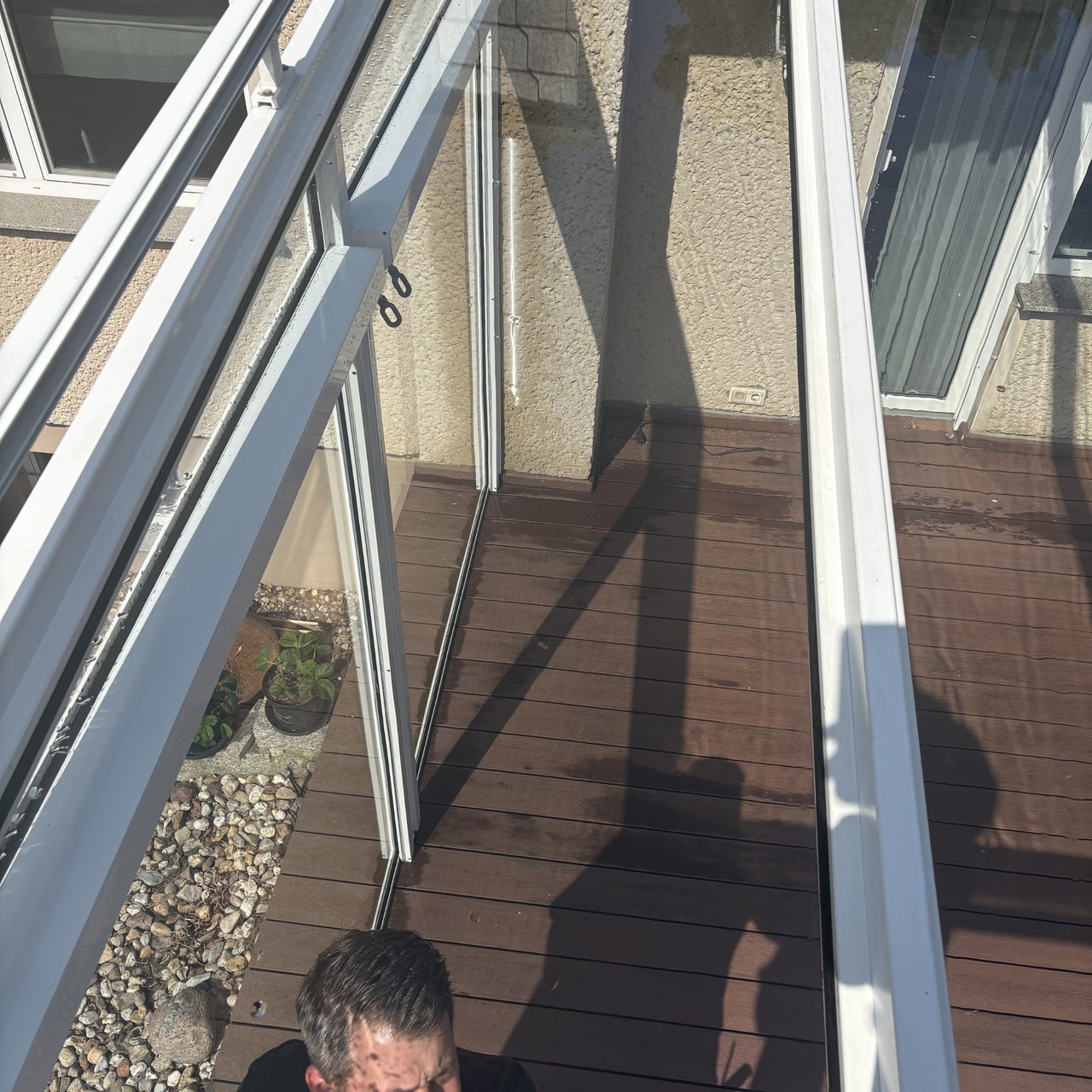 Dunkelbraune Holzterrasse unter einem Glasdach, mit Kiesbeet und Schatten eines Mannes.