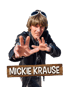Sänger Mickie Krause mit Perücke, Sonnenbrille und Lederjacke vor schwarzem Hintergrund mit Namenszug auf einem Holzschild.