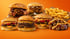 Hände halten einen Burger mit knusprigem Patty, Tomaten, Sprossen und Sauce, der aus einer kleinen blauen Schüssel ragt.