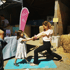 Eine Frau und ein Kind machen gemeinsam Partner-Yoga auf einer blauen Matte vor einem pinken Yoga-Banner und Strohballen.