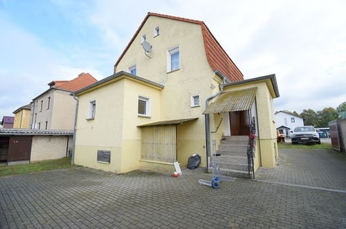 Gelbes Haus mit rotem Ziegeldach, gepflasterter Einfahrt und Außentreppe.