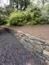 Gartenmauer aus gestapelten Natursteinen trennt einen dunklen Kiesweg von einer bepflanzten Böschung mit grünen Sträuchern.