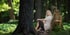 Blonde Frau sitzt barfuß im Wald an einem Baumstumpf, auf dem ein Uhu mit geflecktem Gefieder sitzt.