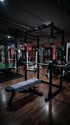 Dunkles Fitnessstudio mit Hantelbank und Langhantel im Power Rack auf Holzboden, andere Geräte im Hintergrund.