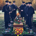 Sechs Bestatter in Uniform stehen um einen Sarg mit Blumenarrangements.