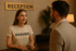 Eine lächelnde junge Frau in einem T-Shirt mit der Aufschrift "OCEVOO" steht hinter einer Rezeption unter einem Schild mit der Aufschrift "RECEPTION" und spricht mit einem Mann im Anzug.