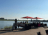 Gäste sitzen unter roten Sonnenschirmen auf einer Terrasse am Flussufer mit grünen Feldern am Horizont.