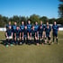 Eine Gruppe von 13 Fußballspielern in blauen Trikots posiert auf einem grünen Rasenfeld unter blauem Himmel.