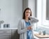 Woman in a kitchen holding a mug while rubbing her neck with a pained expression.
