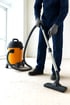 Eine Person in blauer Arbeitskleidung und grauen Handschuhen saugt mit einem orangefarbenen Industriesauger einen schmutzigen Boden.