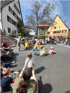 Kinder in bunten Kostümen tanzen auf einem Platz mit Zuschauern und Gebäuden im Hintergrund.