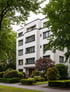 Weißes, modernes Apartmentgebäude mit flachem Dach, umgeben von grünen Bäumen und gepflegten Sträuchern.