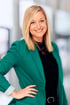 Lächelnde Frau mit blonden Haaren und blau-grauen Augen, die einen grünen Blazer und ein schwarzes Oberteil trägt.