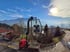 Roter LKW-Kran hebt große Glasscheibe unter blauem Himmel mit Sonne und Bergen.