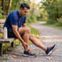 A man in athletic wear sits on a park bench holding his knee and ankle in pain while outdoors on a gravel path.