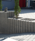 Small cedar trees grow in a stepped concrete retaining wall planter next to a paved stone driveway.