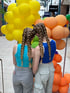 Zwei Frauen mit geflochtenen Zöpfen, von hinten gesehen, stehen vor einem Bogen aus gelben, orangefarbenen und grünen Ballons.