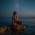 Frau mit roten Locken meditiert im Lotussitz auf einem Felsen im Meer unter einem Sternenhimmel.