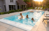 Drei Kinder springen und schwimmen in einem Außenpool vor einem Haus bei Sonnenuntergang.