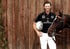 Ein lächelnder Polospieler in Sportkleidung hält einen Helm und steht neben einem braunen Pferd vor einer Holzwand.