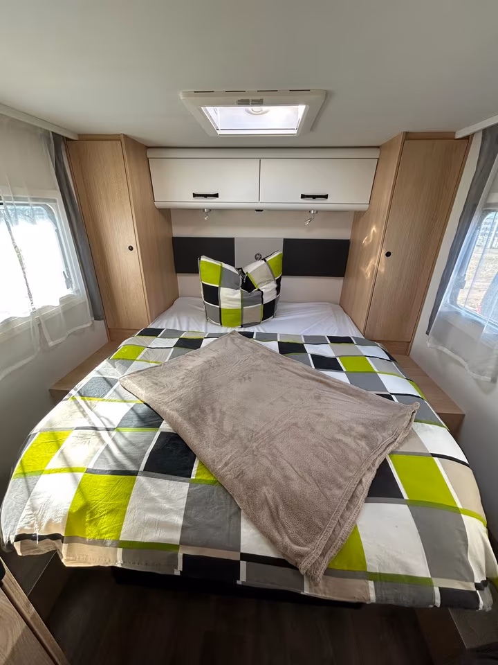 Schlafzimmer im Wohnmobil mit karierter Bettwäsche in Grün, Grau und Schwarz, Holzeinbauschränken und Dachluke.