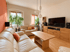 Helles Wohnzimmer mit beigem Ledersofa, Holztisch, TV-Lowboard aus Eiche, Fernseher, Zimmerpflanzen und orangefarbener Wand.
