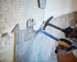 Person mit Arbeitshandschuhen entfernt Fliesen von einer Wand mit einem Hammer und einer Brechstange.